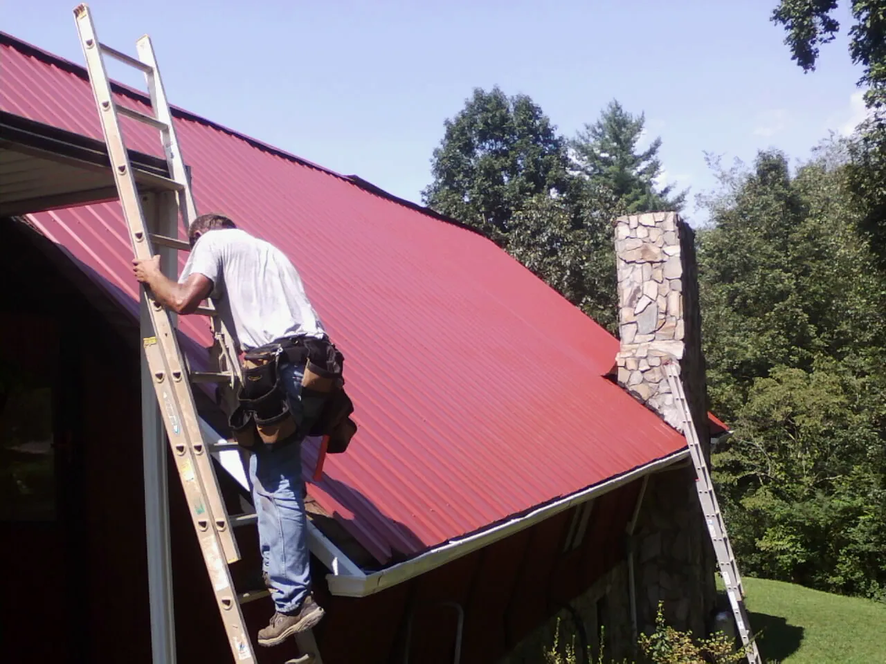 Roofer on a ladder beside a red metal roof and stone chimney — Soffit & Fascia Repair by Roofing Brothers in Saint Louis